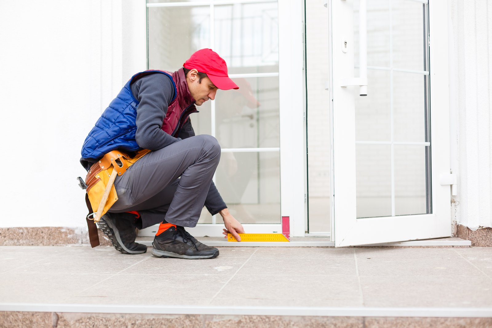 A tradesperson with a tool belt kneeling to measure a residential uPVC door frame