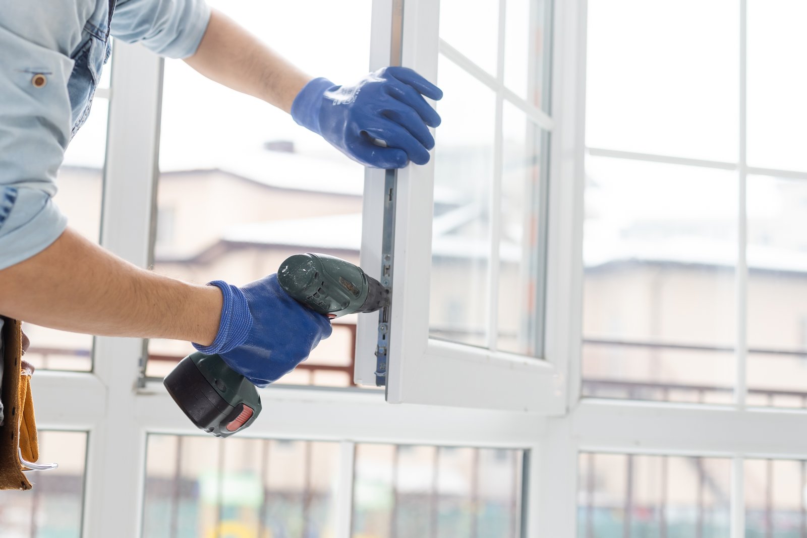 A gloved hand using a drill to fit a lock mechanism into a white window frame