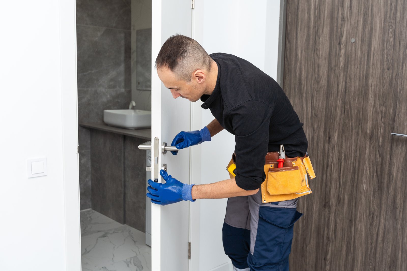 A tradesperson in gloves and a tool belt fitting a handle to an interior door