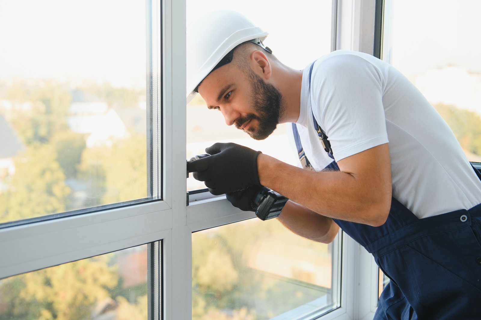 A tradesperson in a hard hat fitting a window lock mechanism with a high-rise view outside