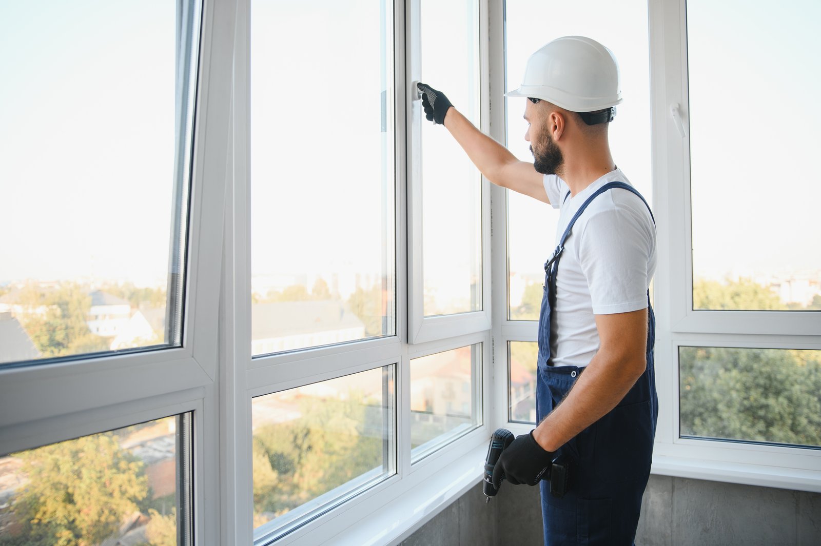 A tradesperson in a hard hat adjusting a window handle indoors with a city view