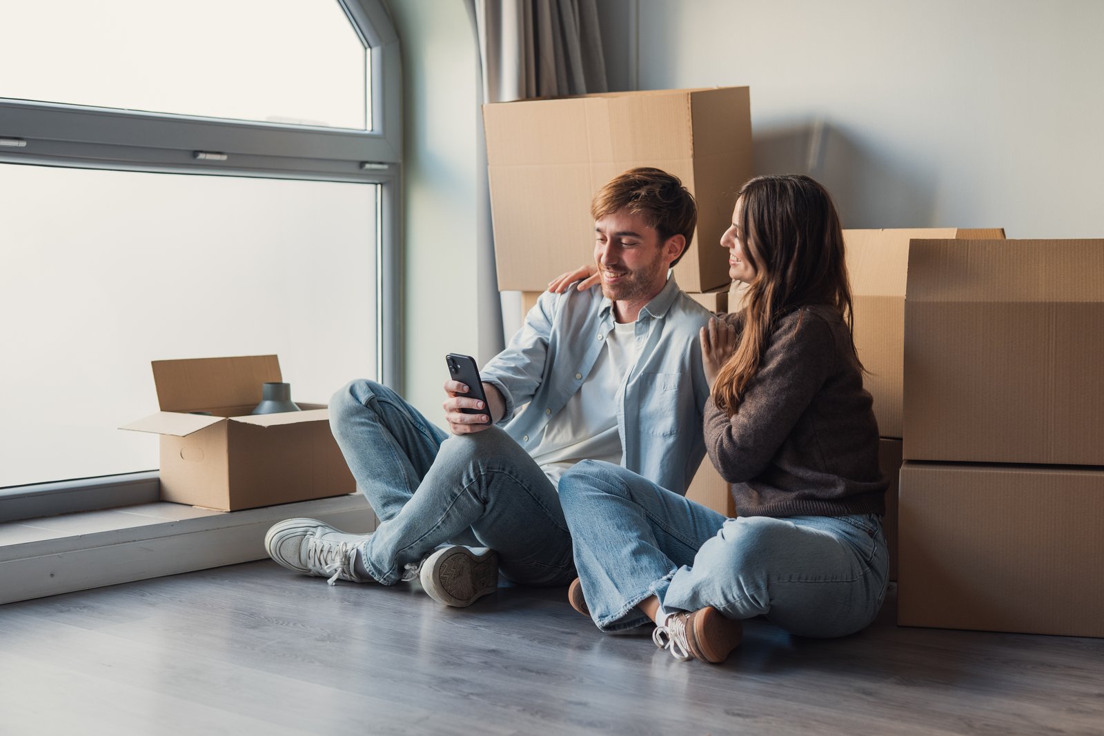 A couple sitting on the floor amid moving boxes, smiling while looking at a phone