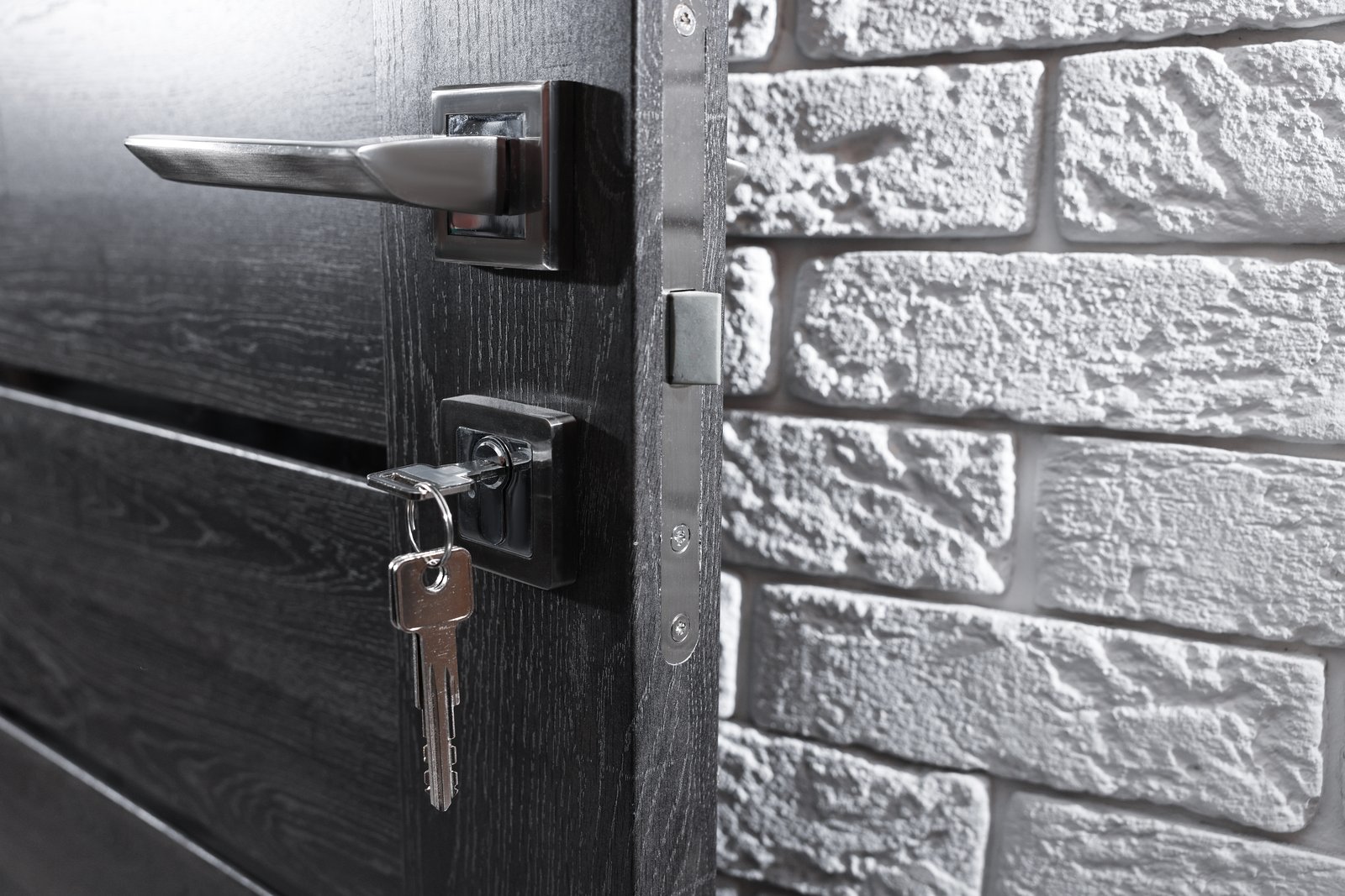 A modern dark door with a lever handle and keys in the lock against a brick wall