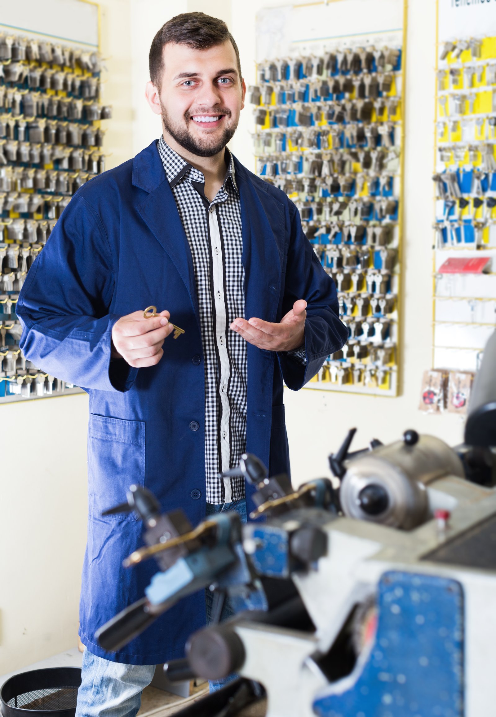 A smiling locksmith holding a key in a workshop lined with key blanks and cutting equipment